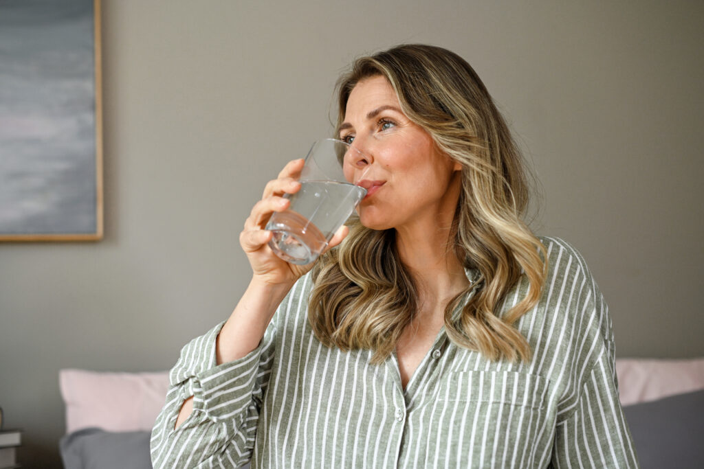 A mature adult woman sits on a bed in her bedroom and drinks water from a glass